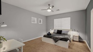 Een slaapkamer met een bed, nachtkastjes, een bureau, een plafondventilator en een raam met jaloezieën.