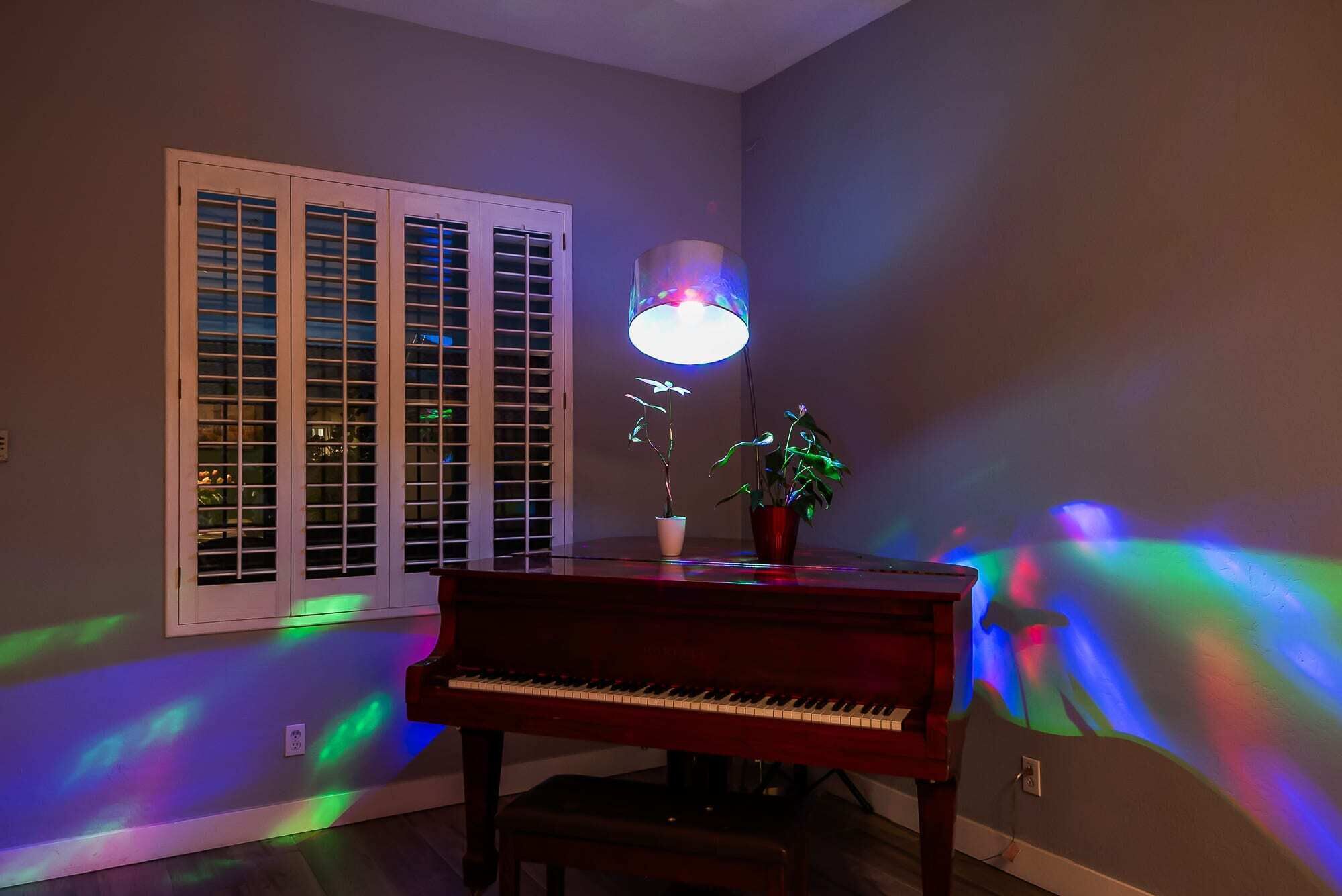 A room with a piano, a window with blinds, and a lamp casting colorful light.