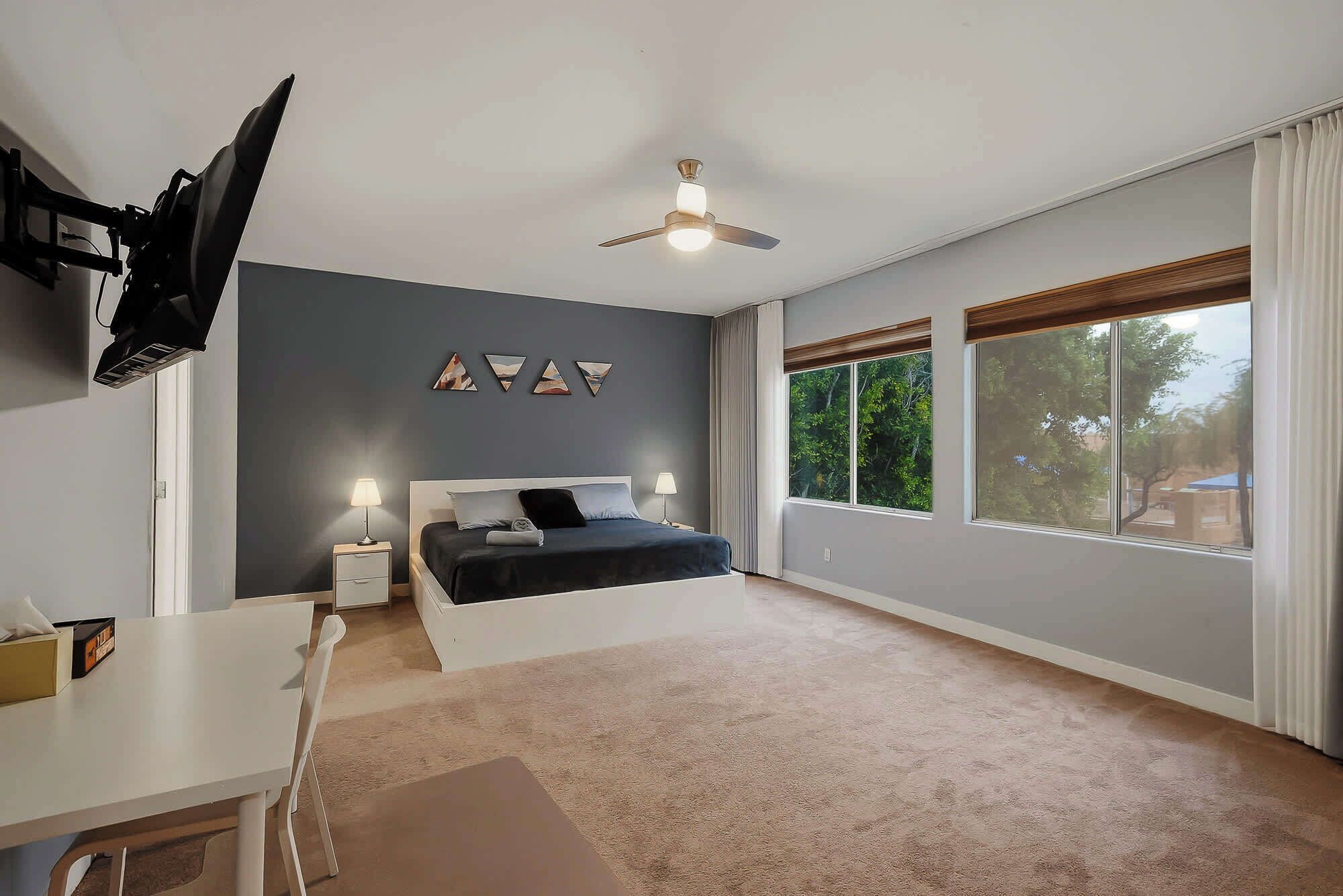 A bedroom with a bed, a desk, a ceiling fan, and large windows.