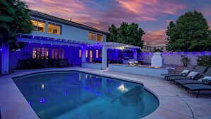 Een zwembad met een terras, een overdekte zithoek en een huis met blauwe sfeerverlichting.