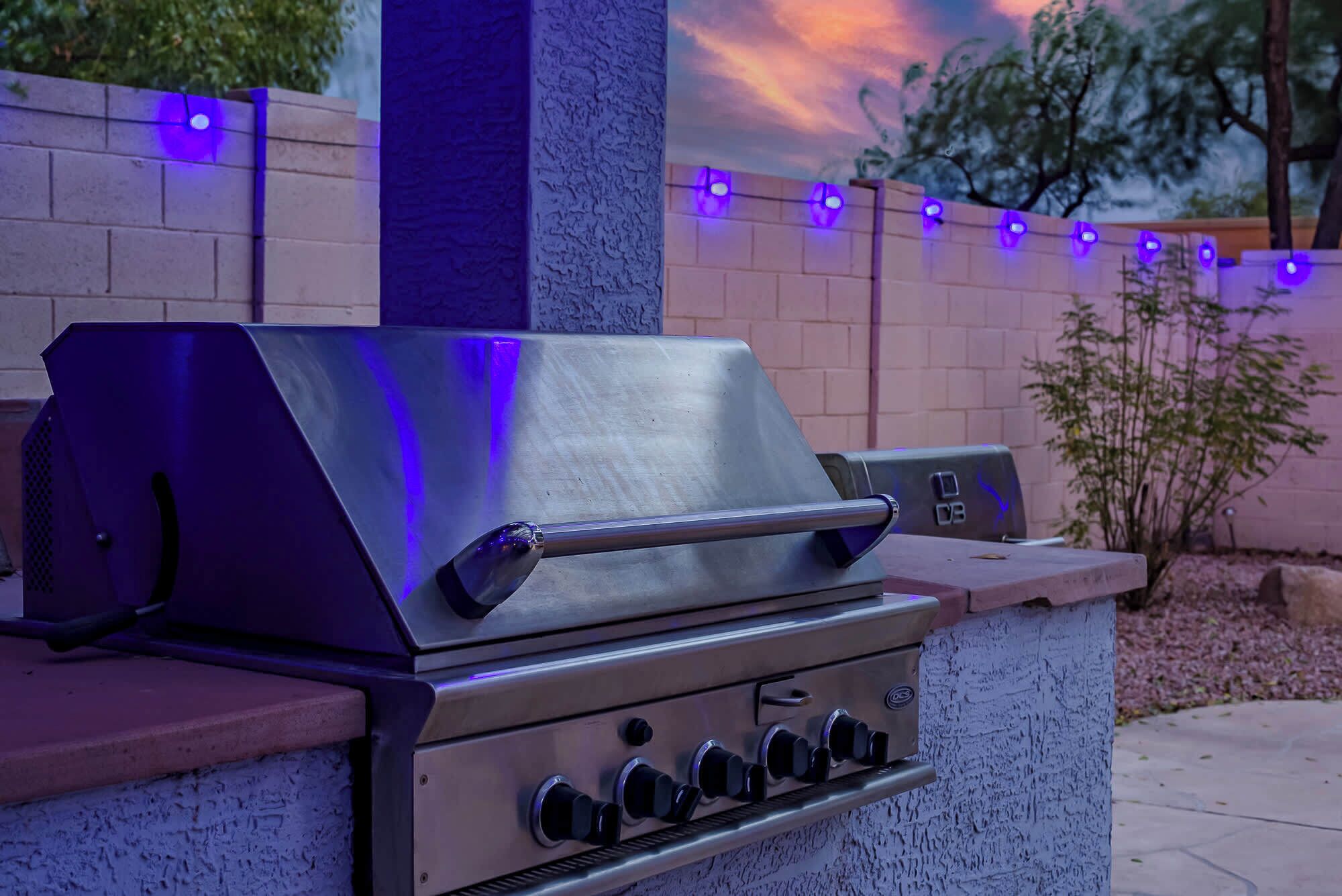 A stainless steel barbecue grill with a control panel, set against a brick wall with a decorative string of lights.
