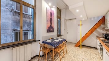 Zona pranzo con tavolo imbandito per quattro persone, sedie di legno, un dipinto raffigurante una torre, una scala e un angolo cottura.