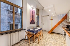 Un coin repas avec une table dressée pour quatre personnes, des chaises en bois, un tableau représentant une tour, un escalier et un espace cuisine.