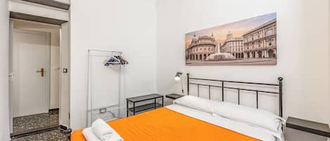 A hotel room with a bed, a nightstand, a lamp, a coat rack, and a painting of a historic building.