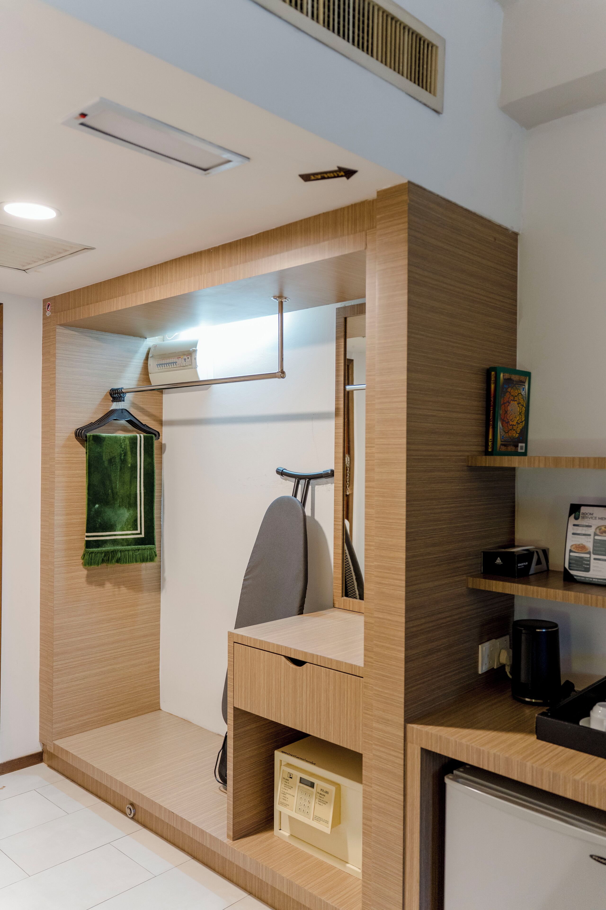 A compact hotel room with a built-in desk, a hanging towel, a small shelf with a book, a coffee machine, and a refrigerator.