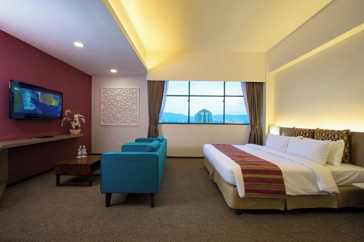 A hotel room with a bed, a desk with a TV, a sofa, and a window with a city view.