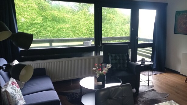 A living room with a sofa, a coffee table, a chair, a TV, and a window with a view of greenery. - House Begonia Room No. 5 (Siegsdorf)