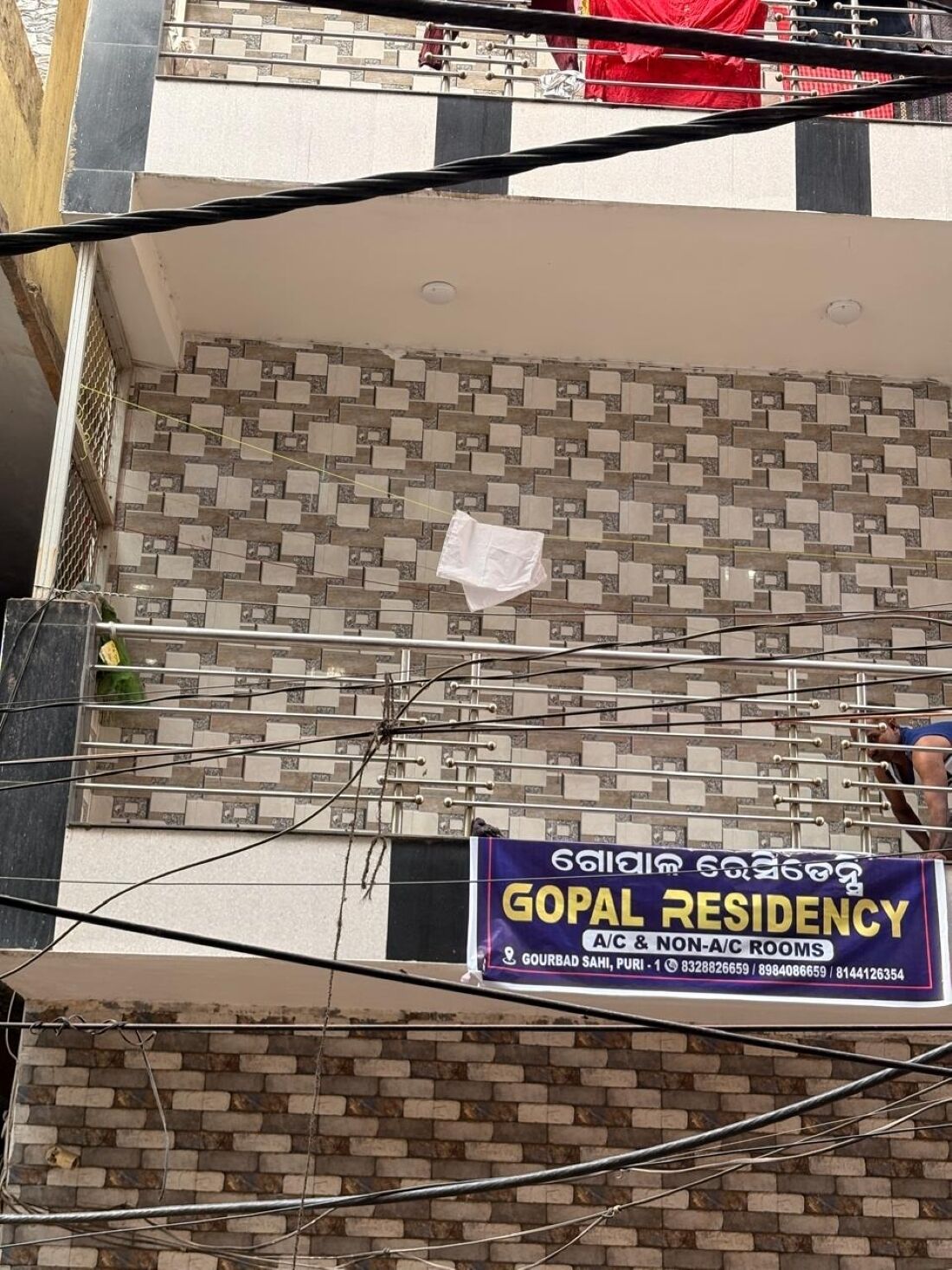 Een gebouw met een betegelde gevel, een bord met Gopal Residency dat kamers met en zonder airconditioning aanbiedt, en een gedeeltelijk zichtbaar balkon met een rode doek.