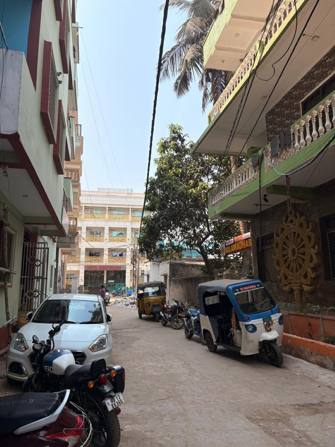 Una calle con vehículos estacionados, incluyendo automóviles y tuk-tuks, y edificios con balcones y rótulos.