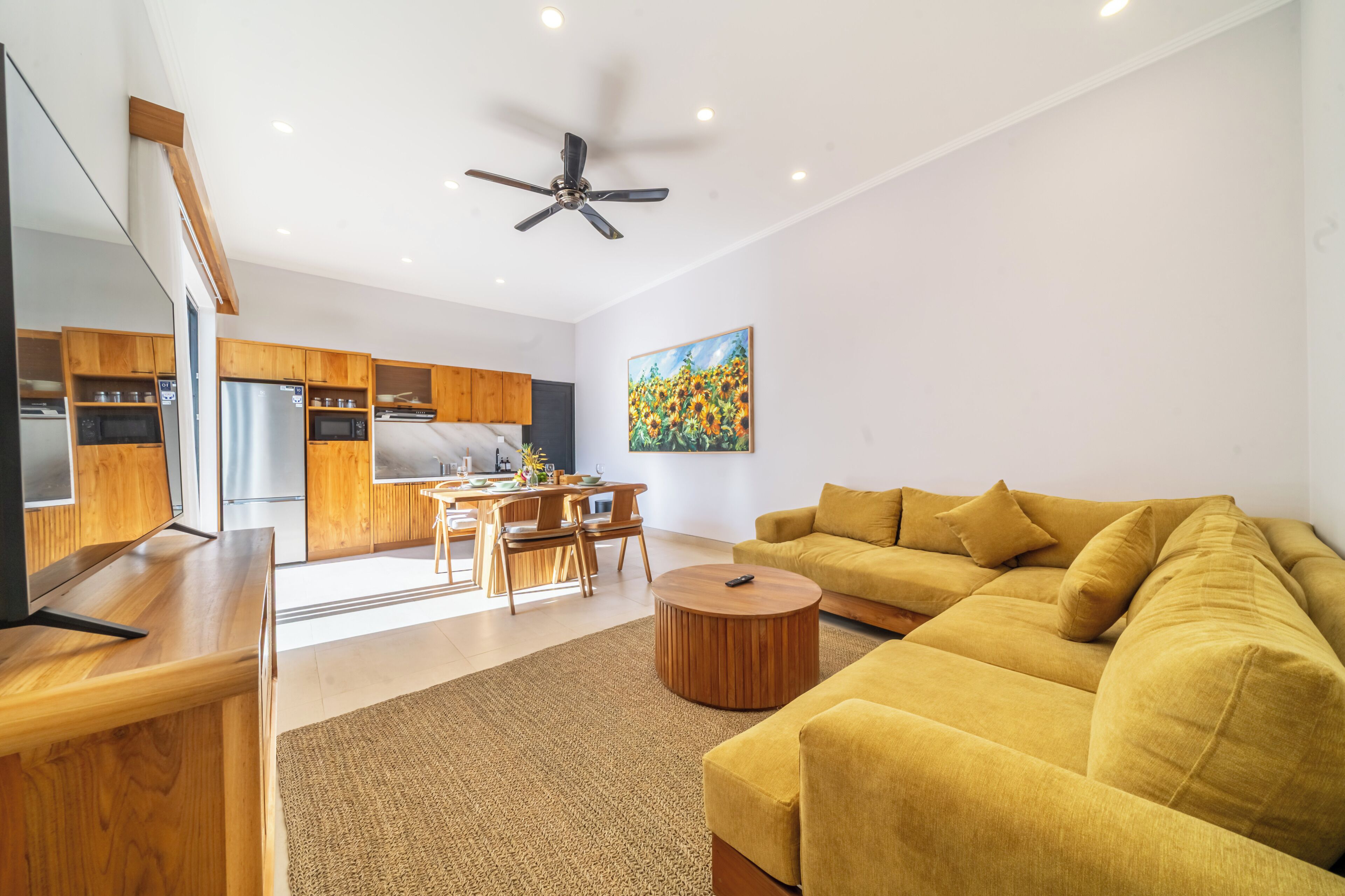 A modern living room with a sofa, a dining area with chairs, a kitchen with wooden cabinets, and a ceiling fan.