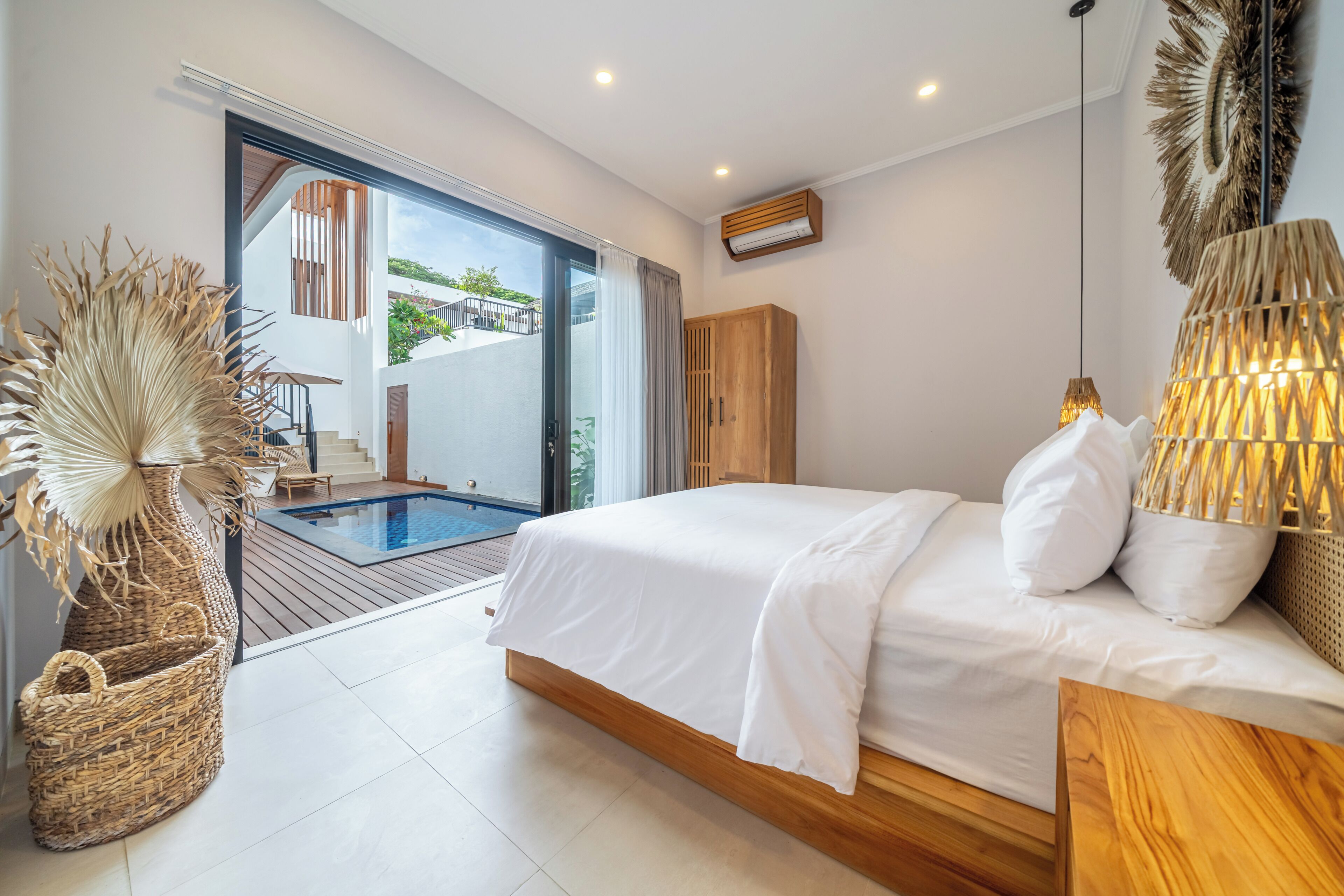 A modern bedroom with a large bed, a wooden wardrobe, a wicker basket, and a view of a pool and outdoor area.