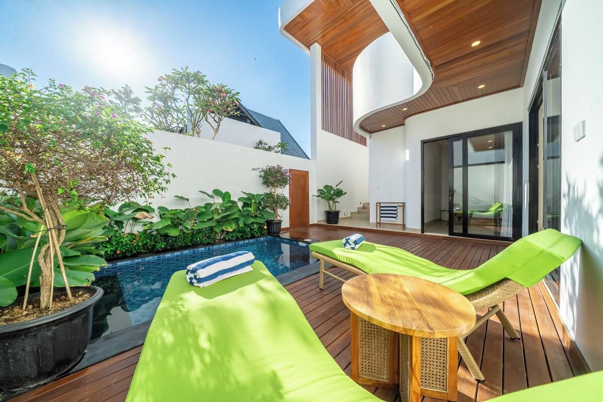 Un área junto a la piscina con sillones de descanso, una mesa de madera y plantas en macetas.