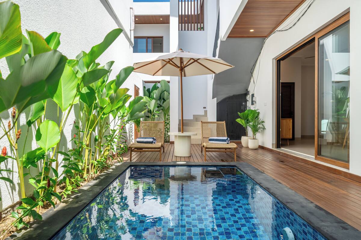 Una piscina con borde de azulejos, rodeada de macetas con plantas y sillas de mimbre.