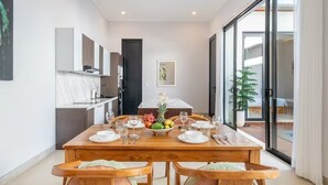 A dining area with a wooden table set for four, a kitchen in the background, and a sliding glass door leading to an outdoor area. - Cassandra Villas - Your Bali Escape (Denpasar)