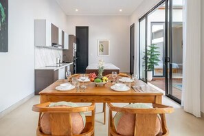 A dining area with a wooden table set for four, a kitchen in the background, and a sliding glass door leading to an outdoor area. - Cassandra Villas - Your Bali Escape (Denpasar)