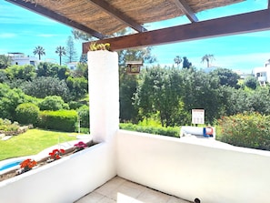 Ein Balkon mit einer weißen Säule, einem Strohdach und Blick auf einen üppigen Garten sowie Gebäude im Hintergrund.