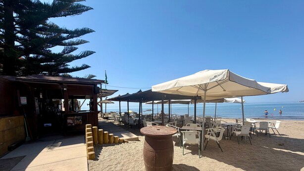 Un café situé en bord de mer, avec des tables à l’extérieur, un tonneau en bois et des parasols.