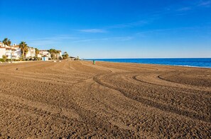 A wide sandy beach with footprints leading to the water, palm trees, and buildings in the background. - Lentiscares 213 Beach & Pool – Torrox Costa (Torrox Costa)
