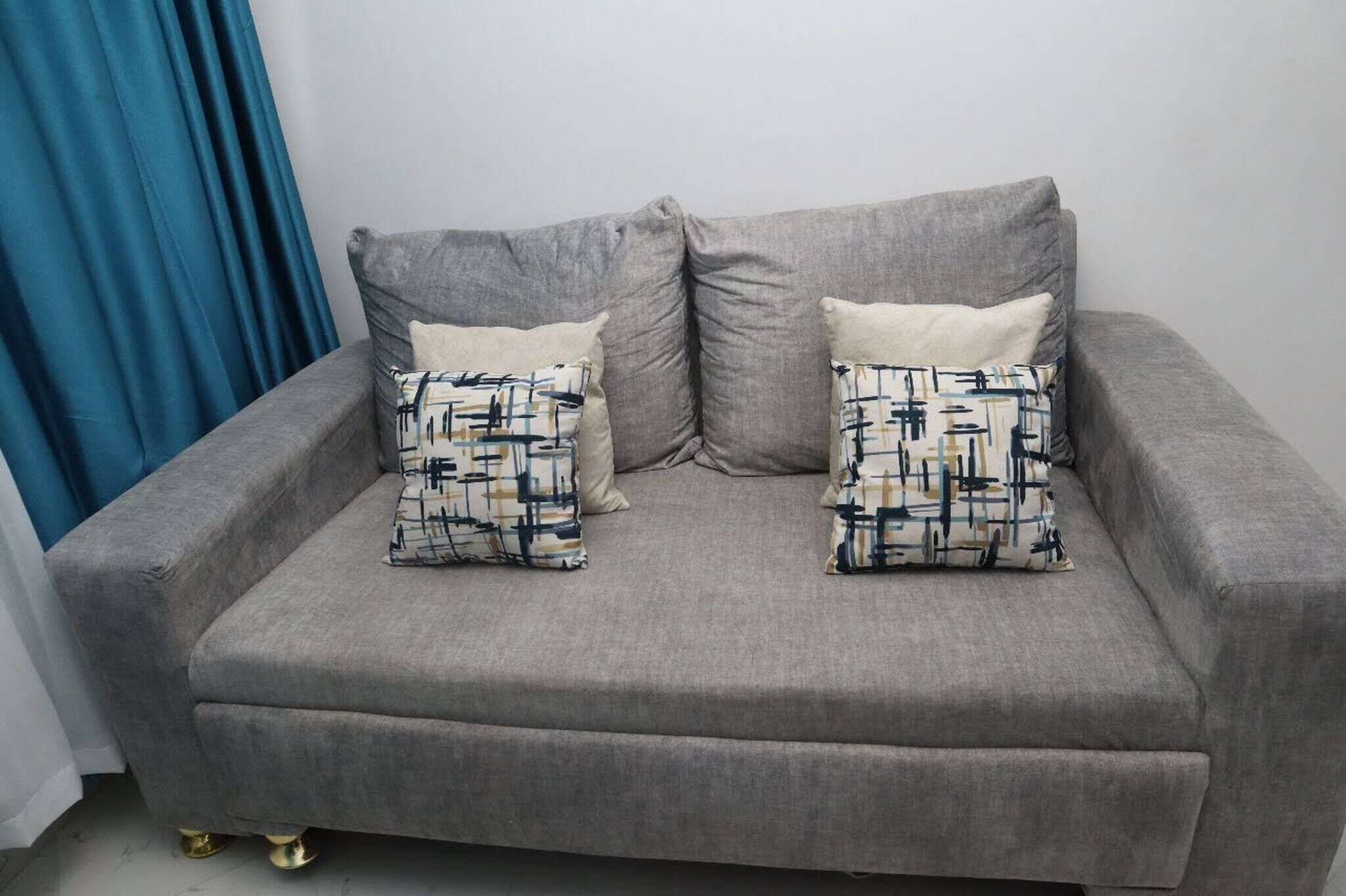 A grey sofa with patterned cushions and gold legs, against a white wall and blue curtain.