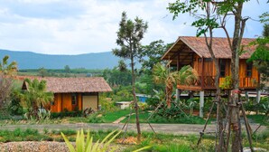 A rustic wooden house with a thatched roof, surrounded by lush greenery and a small pond.