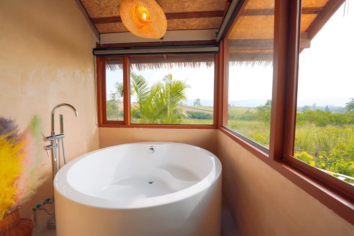 A bathroom with a freestanding bathtub, a window with a view of greenery, and a thatched roof.