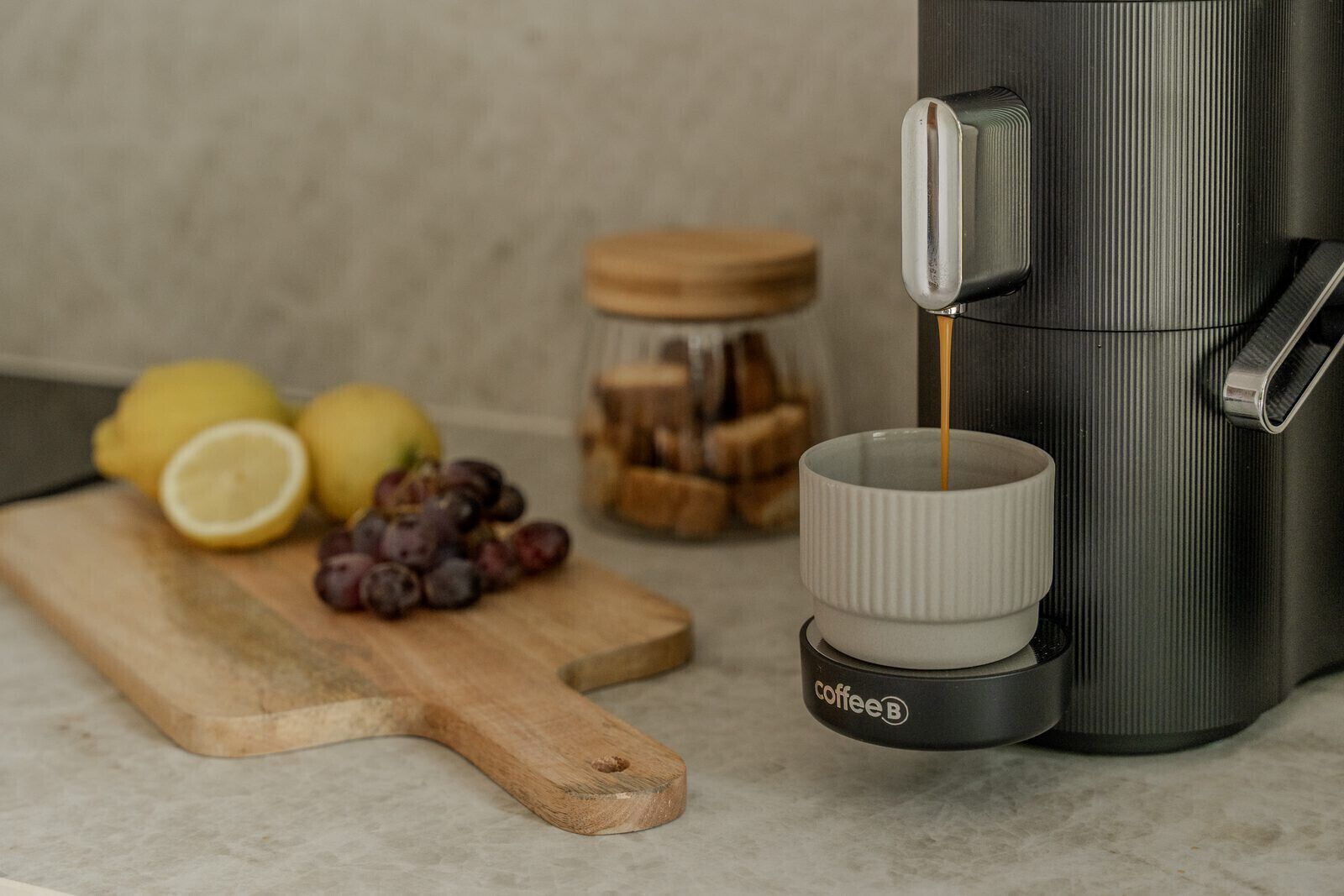 Una cafetera sirviendo café en una taza, una tabla de madera con limones y uvas, y un tarro de miel.