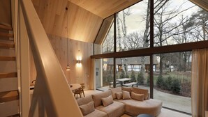 Un intérieur moderne avec un escalier en bois, une grande fenêtre donnant sur les arbres, un canapé d’angle et un coin repas.