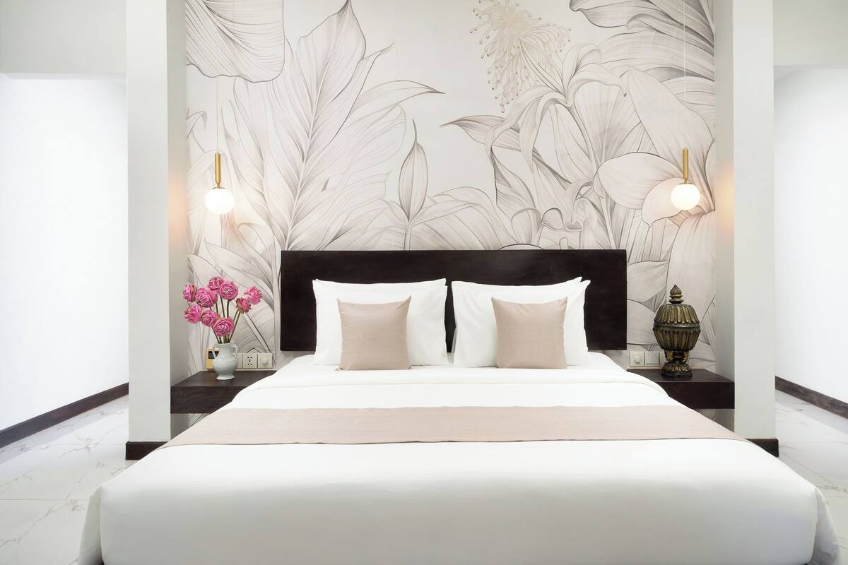 Habitación de hotel con una cama grande, cabecera negra, papel tapiz con motivos florales, dos lámparas de noche y un jarrón con flores rosas.