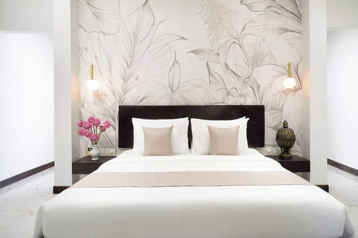 A hotel room with a large bed, a black headboard, a floral wallpaper, two bedside lamps, and a vase with pink flowers.