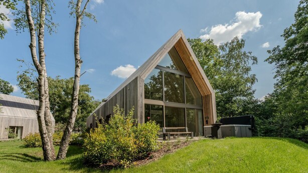 A modern wooden cabin with large windows and a sloped roof, surrounded by lush greenery and trees.