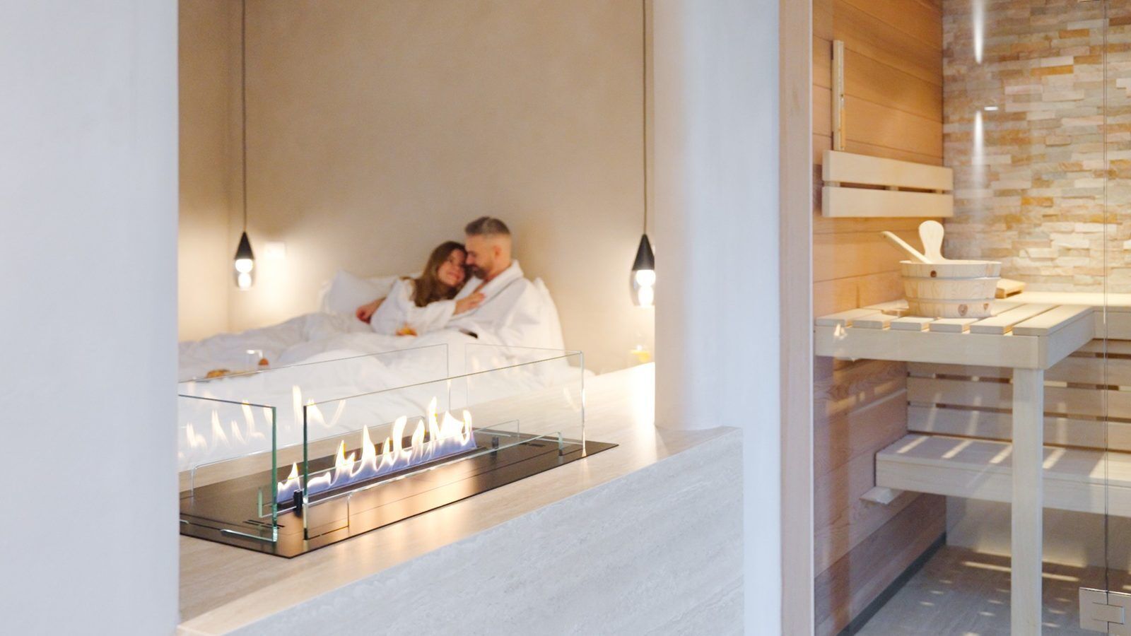 Quarto de hotel moderno com lareira em vidro, cama com roupa de cama branca e parede de madeira com prateleira embutida.