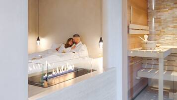 Quarto de hotel moderno com lareira em vidro, cama com roupa de cama branca e parede de madeira com prateleira embutida.