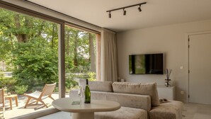 Sala de estar moderna com sofá, mesa redonda pequena, televisão e uma janela grande com vista para vegetação.