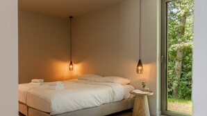 Quarto moderno com uma cama grande, duas luminárias pendentes, uma mesa de cabeceira redonda de madeira e uma janela ampla com vista para área verde.