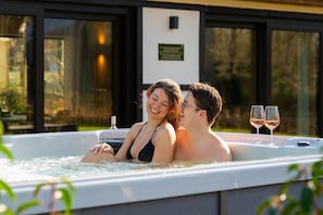 Un couple dans un jacuzzi, avec deux verres de vin, entouré de grandes fenêtres et offrant une vue sur les arbres.