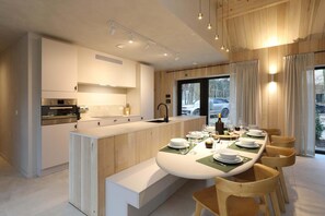 Une cuisine moderne dotée d’appareils électroménagers intégrés, un coin repas avec une longue table et des chaises, et une vue sur l’extérieur grâce à de grandes fenêtres.