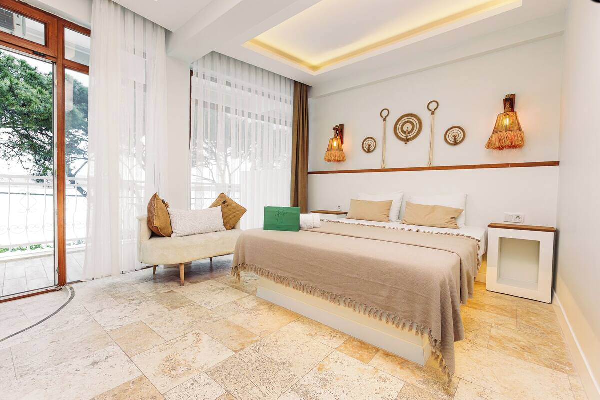 A modern hotel room with a large bed, a sofa, a small table, and a view of the outdoors.