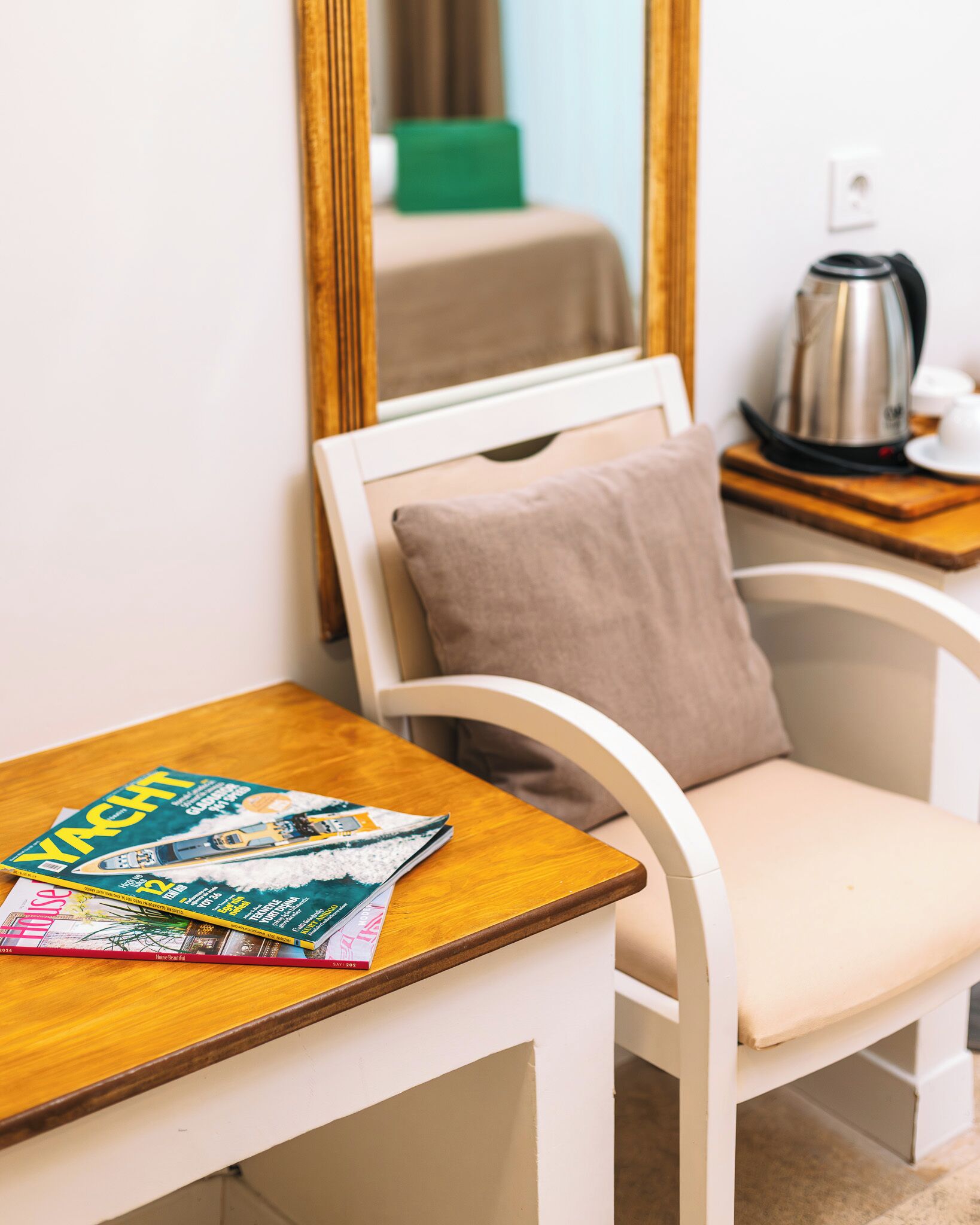 A room with a wooden table, a white chair with a cushion, a magazine, a mirror, a green box, and a kettle on a wooden side table.