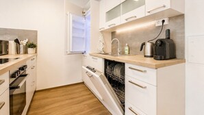 Una cocina moderna con armarios blancos, lavavajillas, cafetera y ventana con persianas.