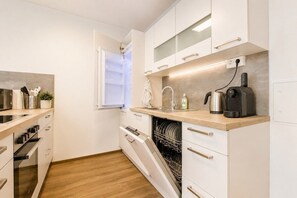 A modern kitchen with white cabinetry, a dishwasher, a coffee maker, and a window with blinds. - Helles City Apartment mit Terrasse (Wien)
