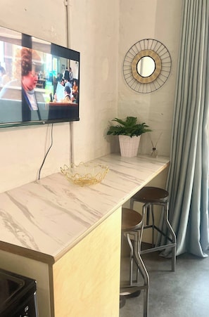 Ein modernes Zimmer mit Marmorplatte, einem an der Wand montierten Fernseher, einem runden Spiegel, einer Topfpflanze und zwei Metallstühlen.