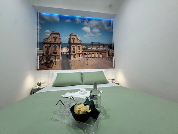 A hotel room with a bed, green bedding, a nightstand with a lamp, a wall-mounted picture of a historical building, and a clear glass tray with snacks and a bottle. - Da Rosalia Apartment: all the comforts in the heart of Palermo's historic center (Palermo)