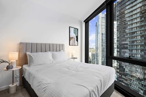 A modern hotel room with a large bed, a nightstand, a lamp, a framed picture, and a view of high-rise buildings. - Southbank 2BR Apt | Near Crown Casino Pool & Gym (Southbank)