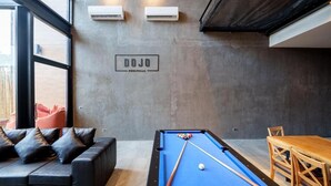 A pool table with a blue felt surface, a sofa with cushions, a wooden table with chairs, and a sign on the wall. - Dojo poolvilla beach resort (Sattahip)