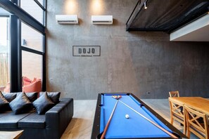 Ein Billardtisch mit blauer Spieloberfläche, eine Couch mit Kissen, ein Holztisch mit Stühlen und ein Schild an der Wand.