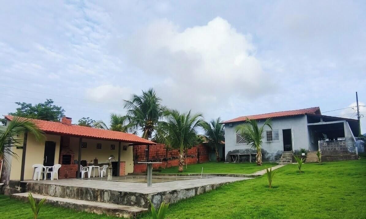Uma casa com telhado de telhas vermelhas, um alpendre com cadeiras e um gramado com palmeiras.