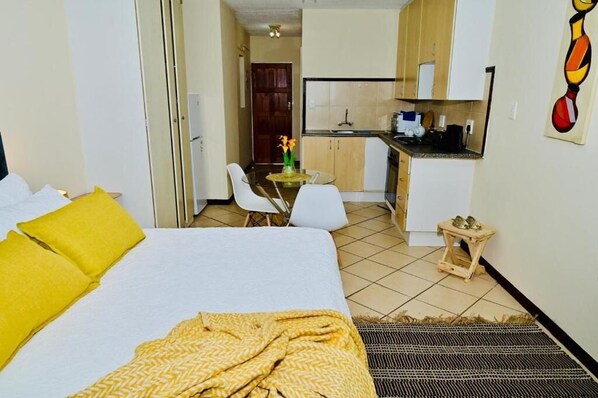 A hotel room with a bed, a small table with chairs, a kitchenette, and a door leading to another room.