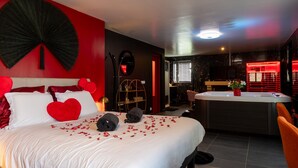 Ein Hotelzimmer mit Bett, roter Wand, großem schwarzen Ventilator, rundem Spiegel, rotem herzförmigen Kissen, roter Blumenvase, Whirlpool und kleinem Tisch mit Stuhl.