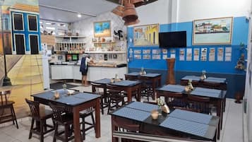 Restaurante com várias mesas e cadeiras, balcão com um atendente na frente e paredes decoradas com quadros e cardápios.