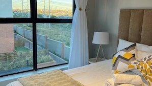 Ein Hotelzimmer mit großem Fenster, einem Bett mit flauschiger Decke, zwei Handtüchern, einer Nachttischlampe und Blick auf eine Stadtlandschaft.